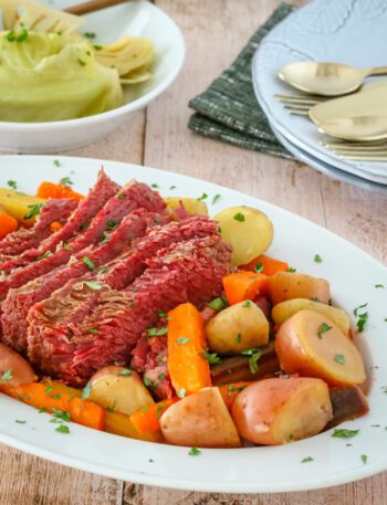corned beef on a platter with potatoes, carrots, and cabbage on a plate in the background
