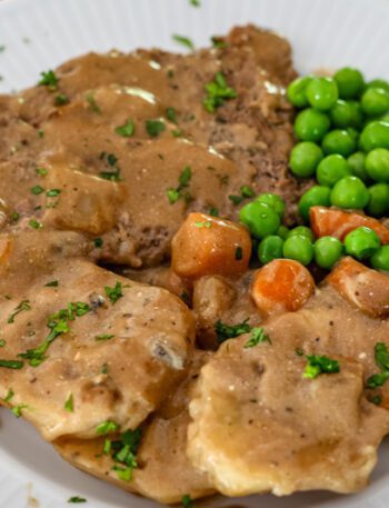 cube steak with mushrooms and gravy shown on a plate with potatoes, peas, and carrots